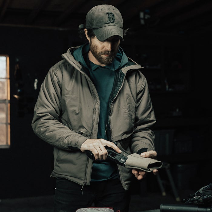Photograph of a man in a taupe jacket and Boston Red Sox baseball cap cleaning a firearm in a dimly lit garage with a window and dark wood shelving visible