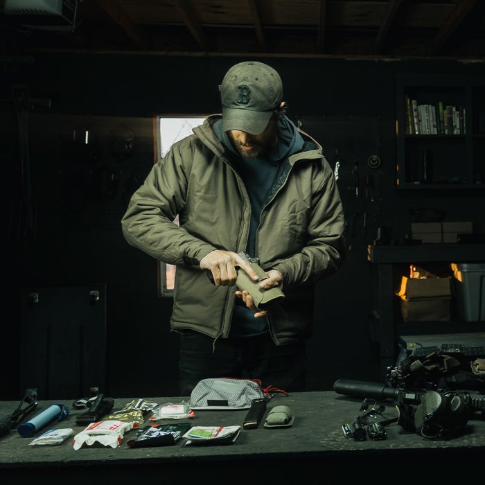 Photography unknown artist a man in an olive green jacket and Boston Red Sox cap prepares a firearm in a dimly lit workshop with a wood burning stove and various survival gear visible including a gray mesh bag and a black knife