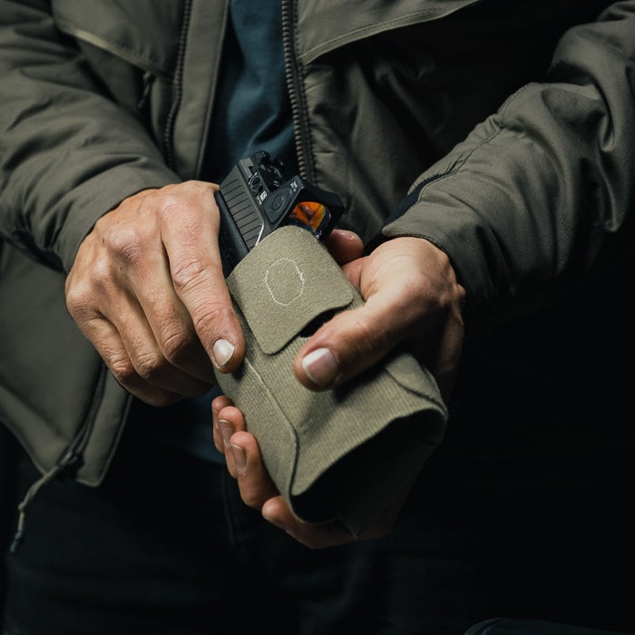 Photograph of a person's hands holding a tan fabric pistol holster containing a black handgun with an orange sight a subtle beige patch and visible stitching