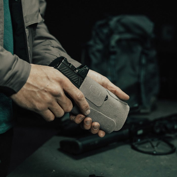 Photograph showing a person holding a black handgun in a gray fabric holster with a textured surface and red sight near a dark green backpack and other firearms on a dark surface