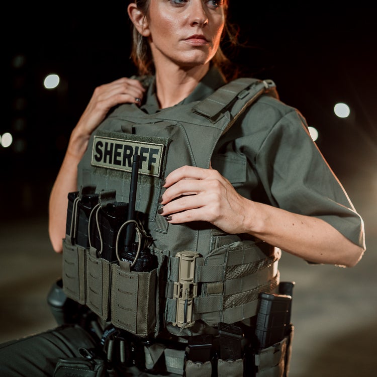 Photography Female Sheriff nighttime outdoor setting wearing a tactical vest with Sheriff patch and radio dark-toned olive-green vest and dark nail polish.