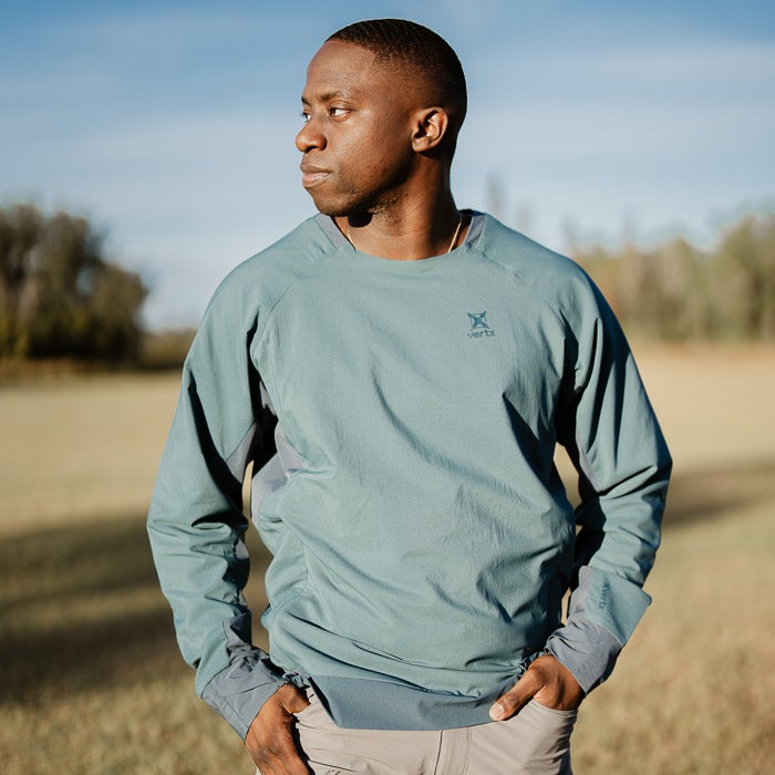 Photograph of a man wearing a teal Vertex long-sleeve shirt outdoors in a field with trees in the background showing the shirt's unique raglan sleeves and subtle logo detail