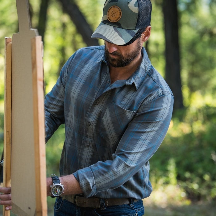Photograph of a man outdoors constructing a wooden frame wearing a blue plaid shirt a camouflage hat and a silver watch in a wooded area