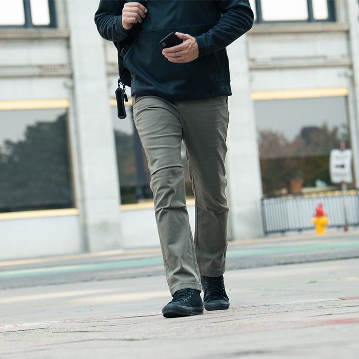 A photograph showing a man walking down a city street wearing olive green pants and black shoes near a fire hydrant and a building with large windows