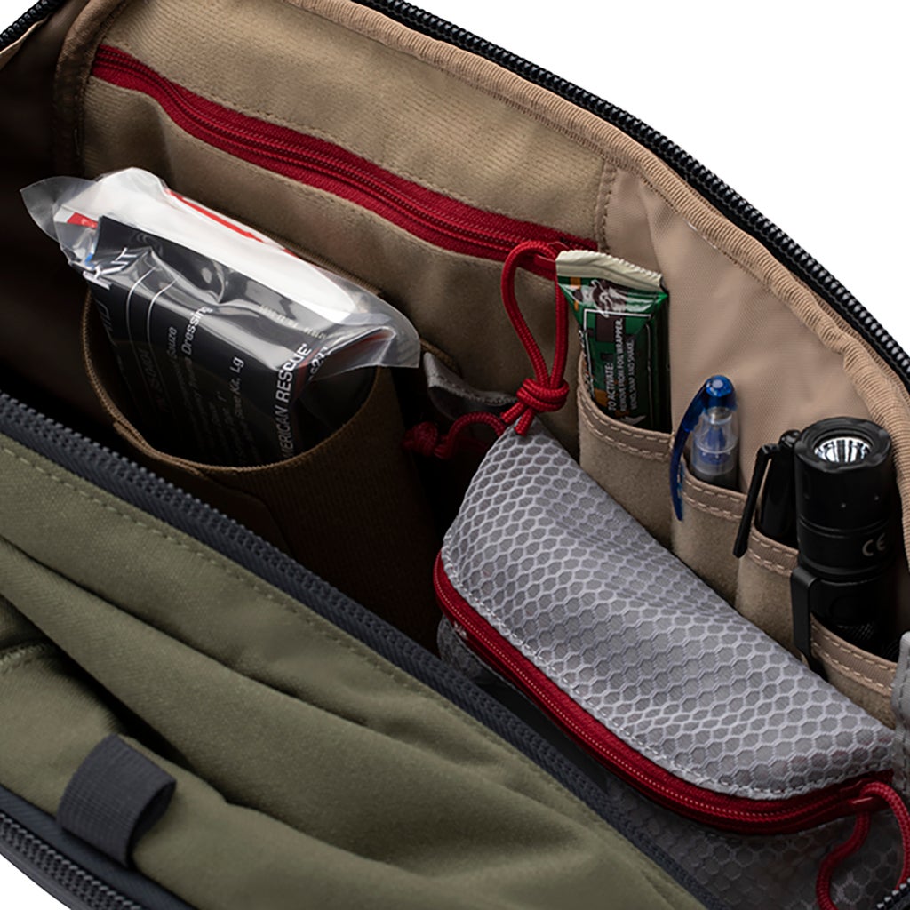 Product Photography: Interior view of a green and tan bag featuring an organized array of items including a first aid kit, energy bar, pens, and flashlight, with red zipper accents and grey mesh pouch.