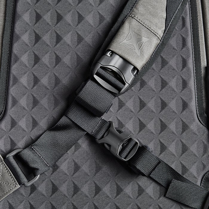 Close-up photograph of a Peak Design backpack detail showing adjustable straps, grey textured fabric and black plastic buckles