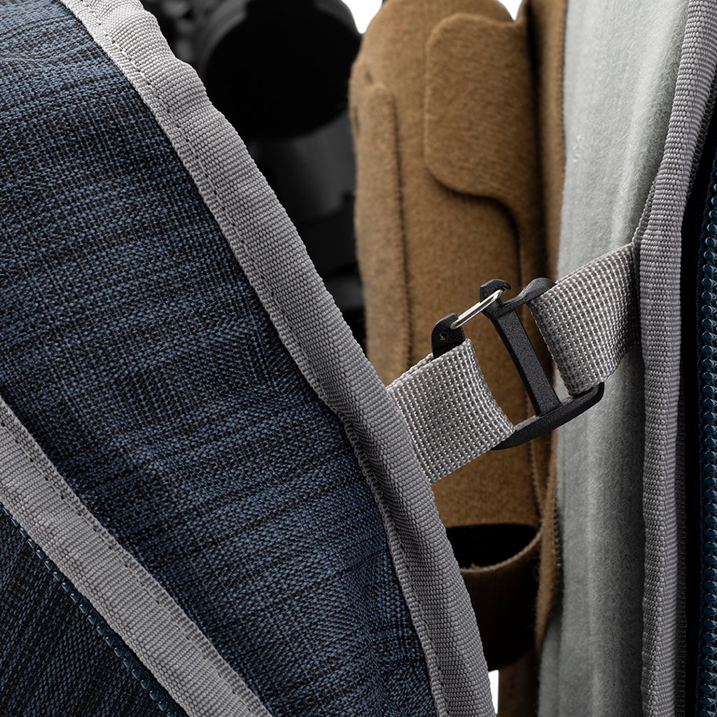 Close-up product shot of a dark blue backpack with gray accents showing a detail of its adjustable strap and plastic buckle featuring a textured fabric and contrasting teal zipper pull
