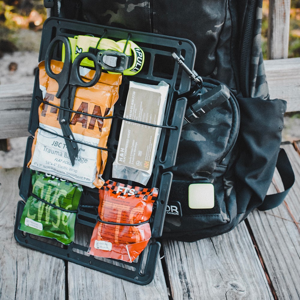 Photograph of a black tactical medical pouch attached to a backpack on a wooden surface showing trauma shears, bandages, and other first-aid supplies in bright orange, green, and yellow packaging.