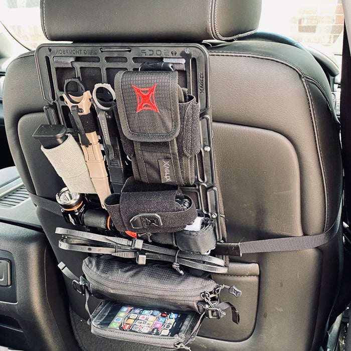 Photograph of a vehicle's backseat organization system showing various tactical gear including a flashlight, knife, and smartphone in a black pouch attached to the headrest with a red X logo and black straps