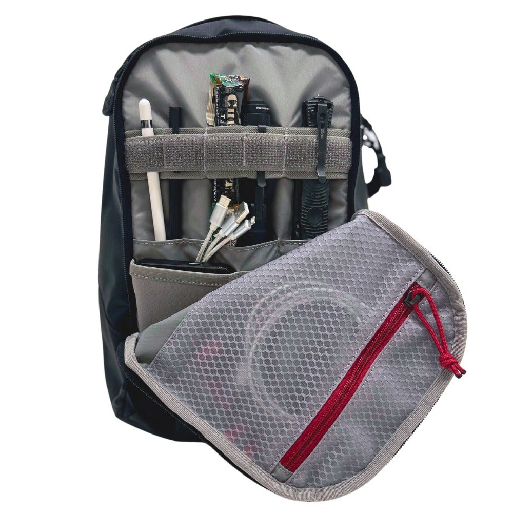 Product photography showing the interior of a black EDC bag with gray organizational pouches, a red zipper, and various tech accessories including a smartphone, charging cables, and a stylus.