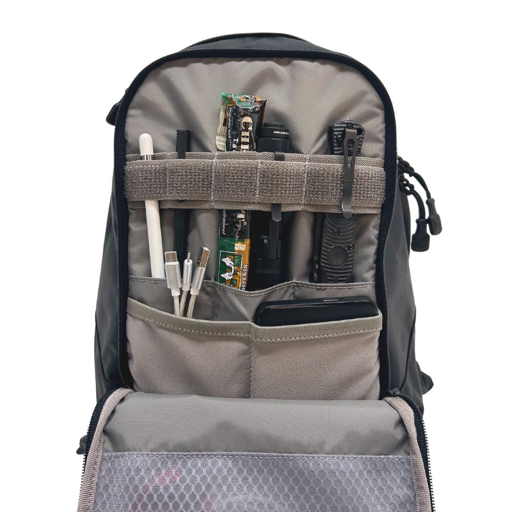 Photograph of a black backpack's interior showing organized pockets with a smartphone, charging cables, pens, a flashlight, and energy bar visible alongside molle webbing and other tactical gear
