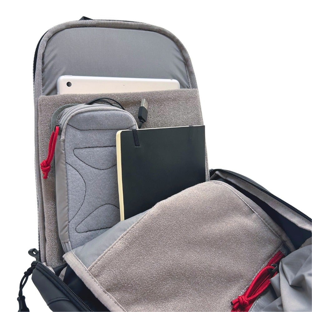 Product shot showing the interior of a black backpack with gray lining containing a tablet, notebook, small pouch, and USB cable featuring red accents and soft gray fabric