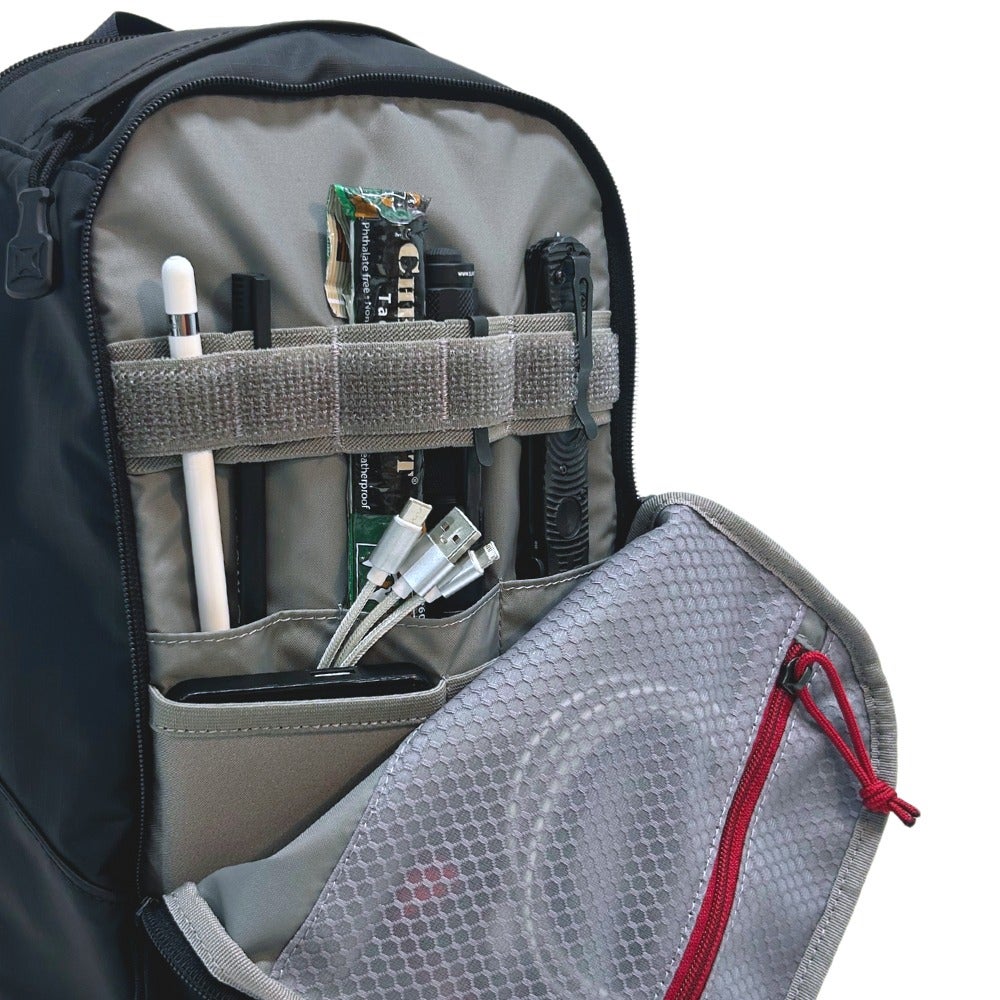 Product photography showing the organized interior of a black backpack with gray compartments featuring a flashlight, charging cables, and a stylus against a white background
