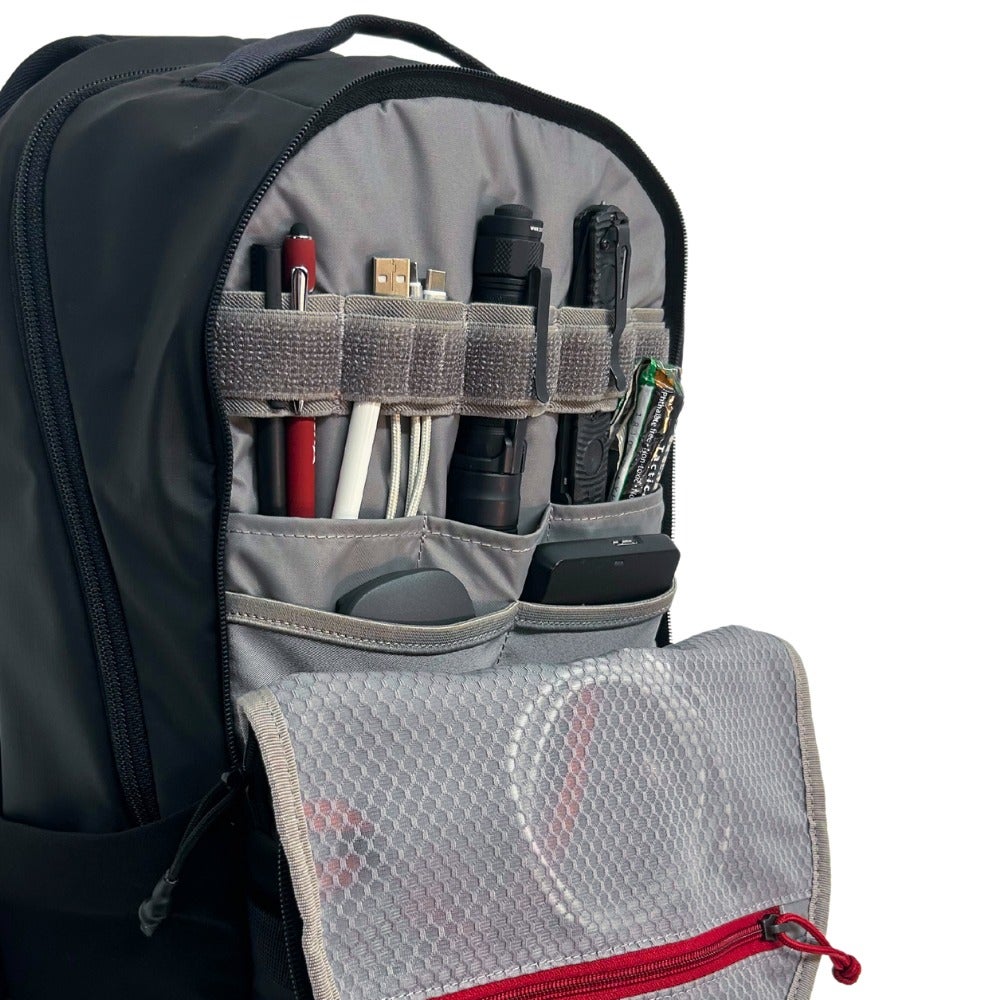 Product shot showing the organized interior of a black backpack with grey compartments, featuring pens, cables, a flashlight, and a knife alongside a red zipper and mesh pocket.