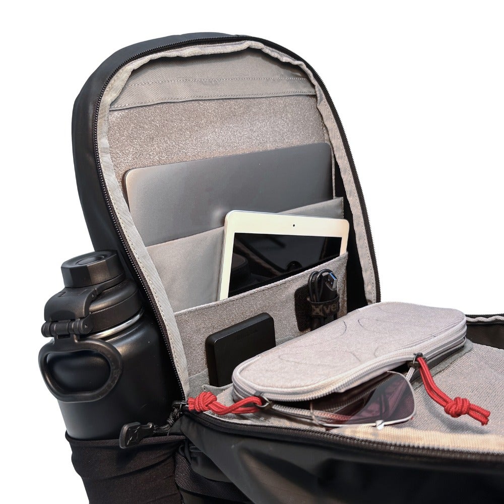 Photograph of a black backpack interior showing a laptop tablet water bottle and sunglasses with grey organizational pockets and red drawstring accents