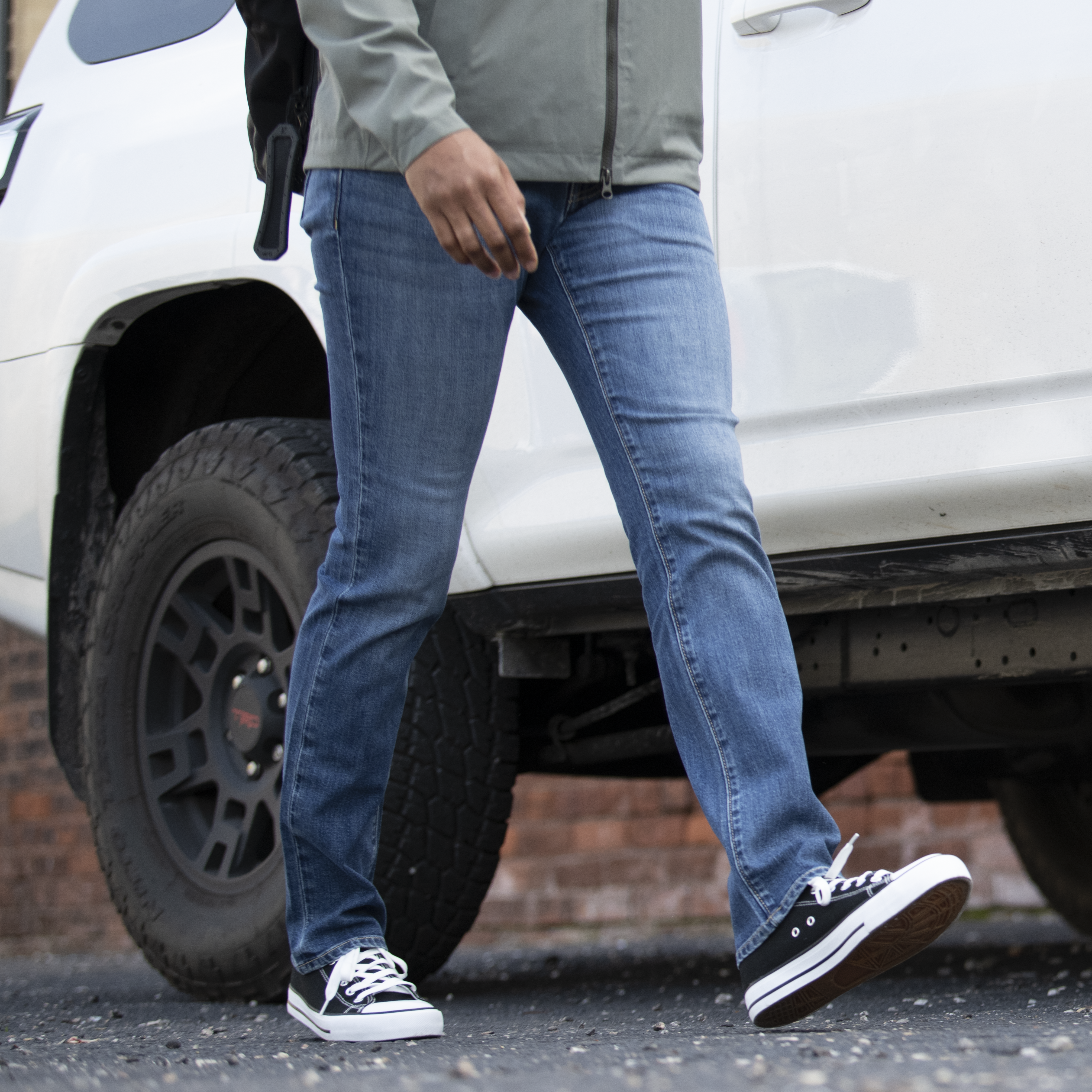 Photograph person walking near a white SUV with black tires and rims next to a brick wall wearing blue jeans and black canvas shoes with white soles and laces