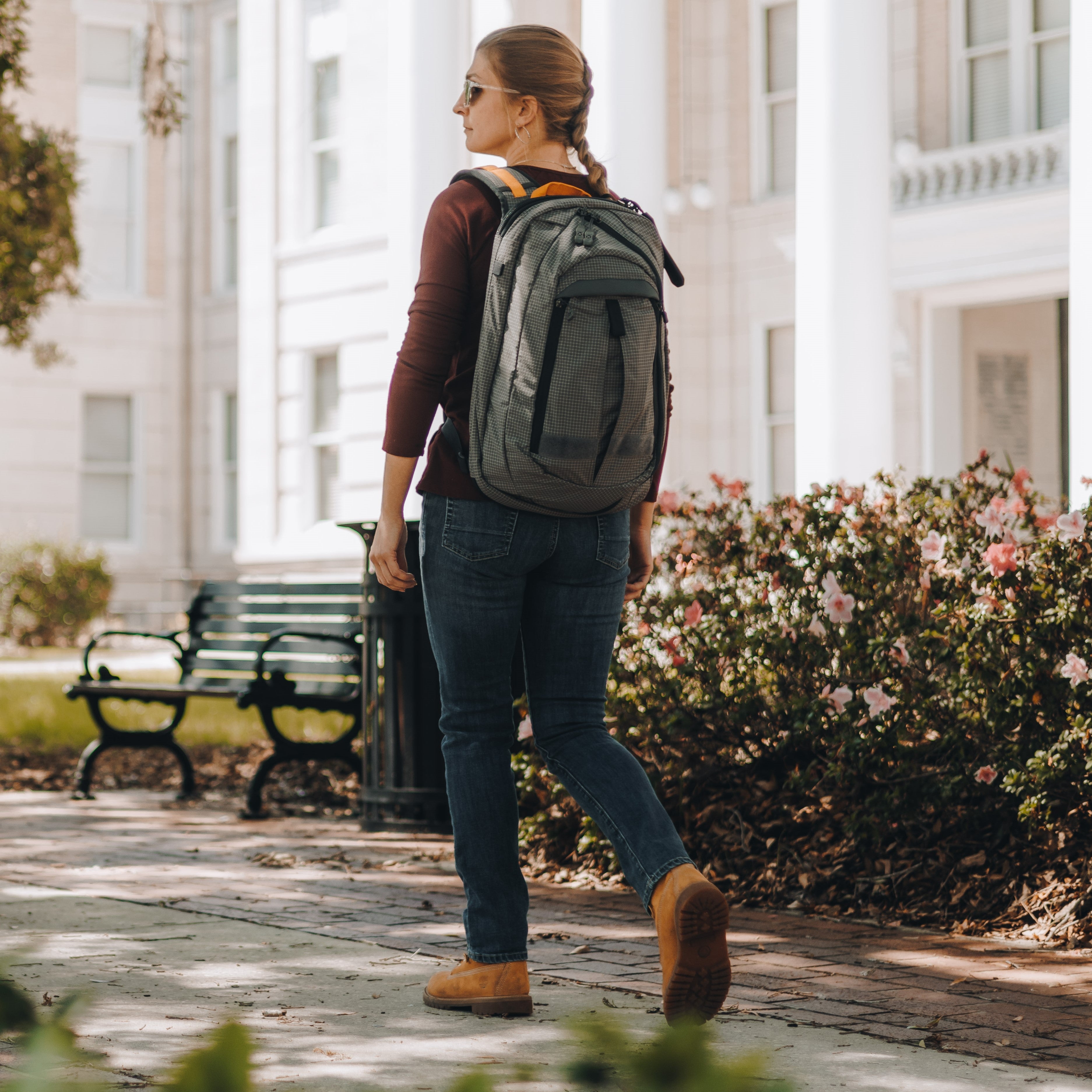 Photography, unknown artist a woman with a grey backpack walks past a white building with columns and pink flowers on a brick walkway wearing brown boots and sunglasses