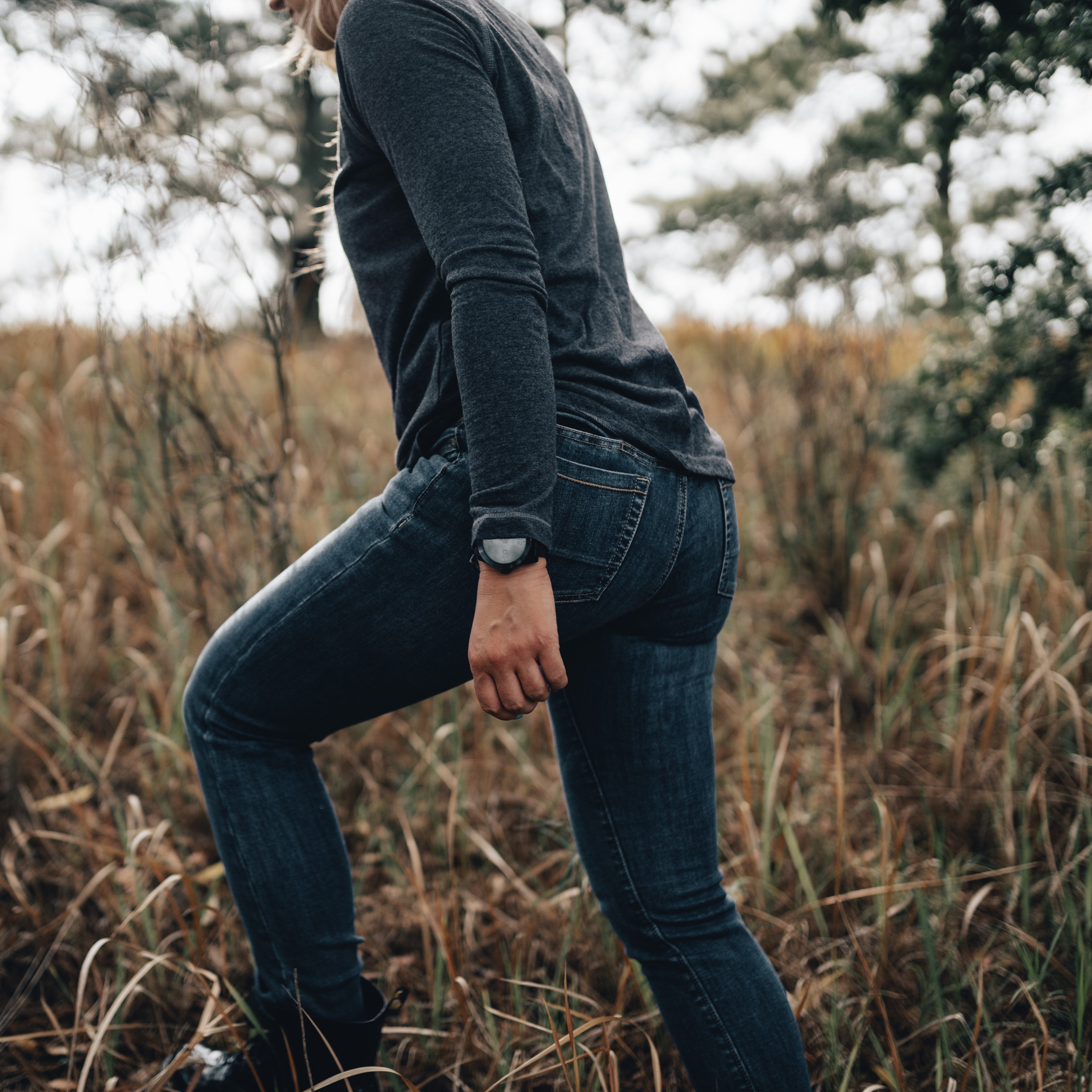Photograph of a person wearing dark gray long sleeves and blue jeans walking through tall, dry grass with a dark-colored watch and a hint of teal nail polish visible.