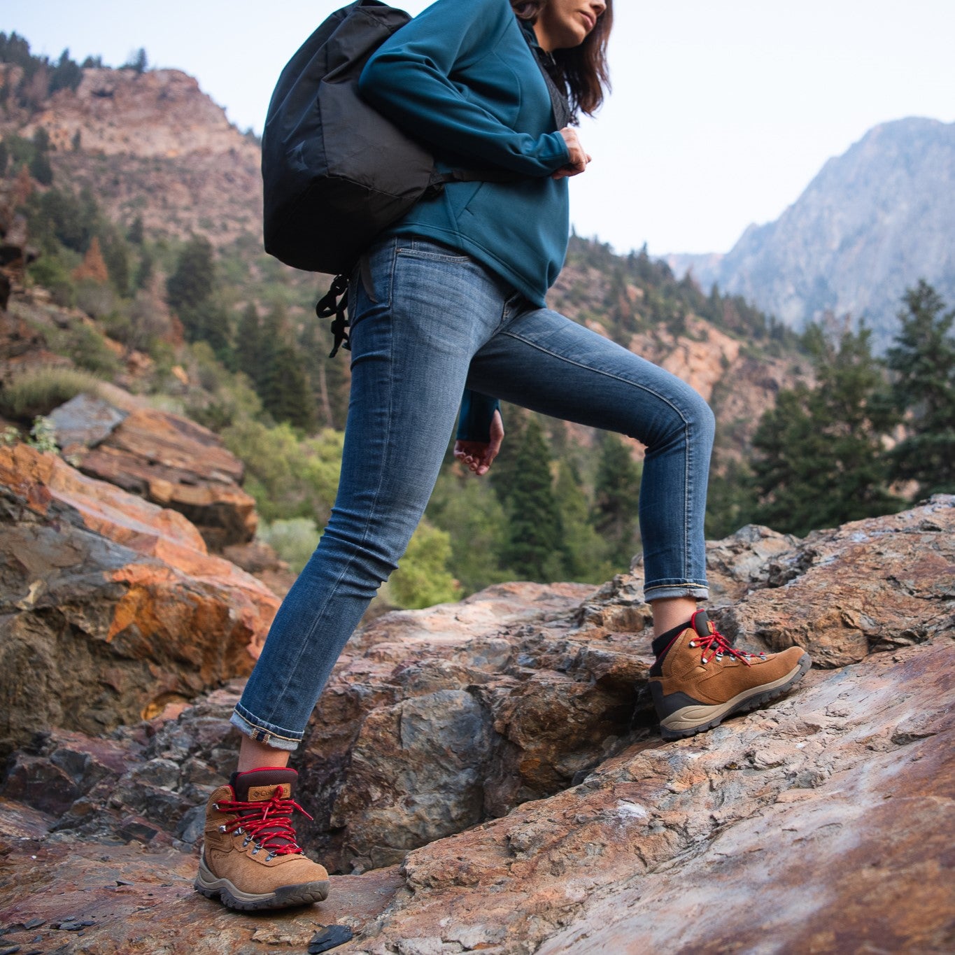 Photograph hiker in teal shirt and jeans hiking rocky mountain trail with red laces on brown boots and a black backpack amidst towering mountains and evergreen trees