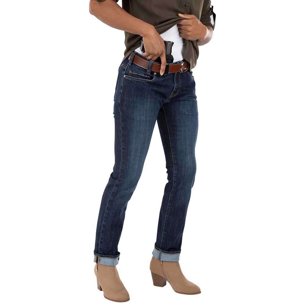 Studio photograph of a person wearing dark-wash jeans a brown belt and tan ankle boots concealing a handgun in a waistband holster against a white background