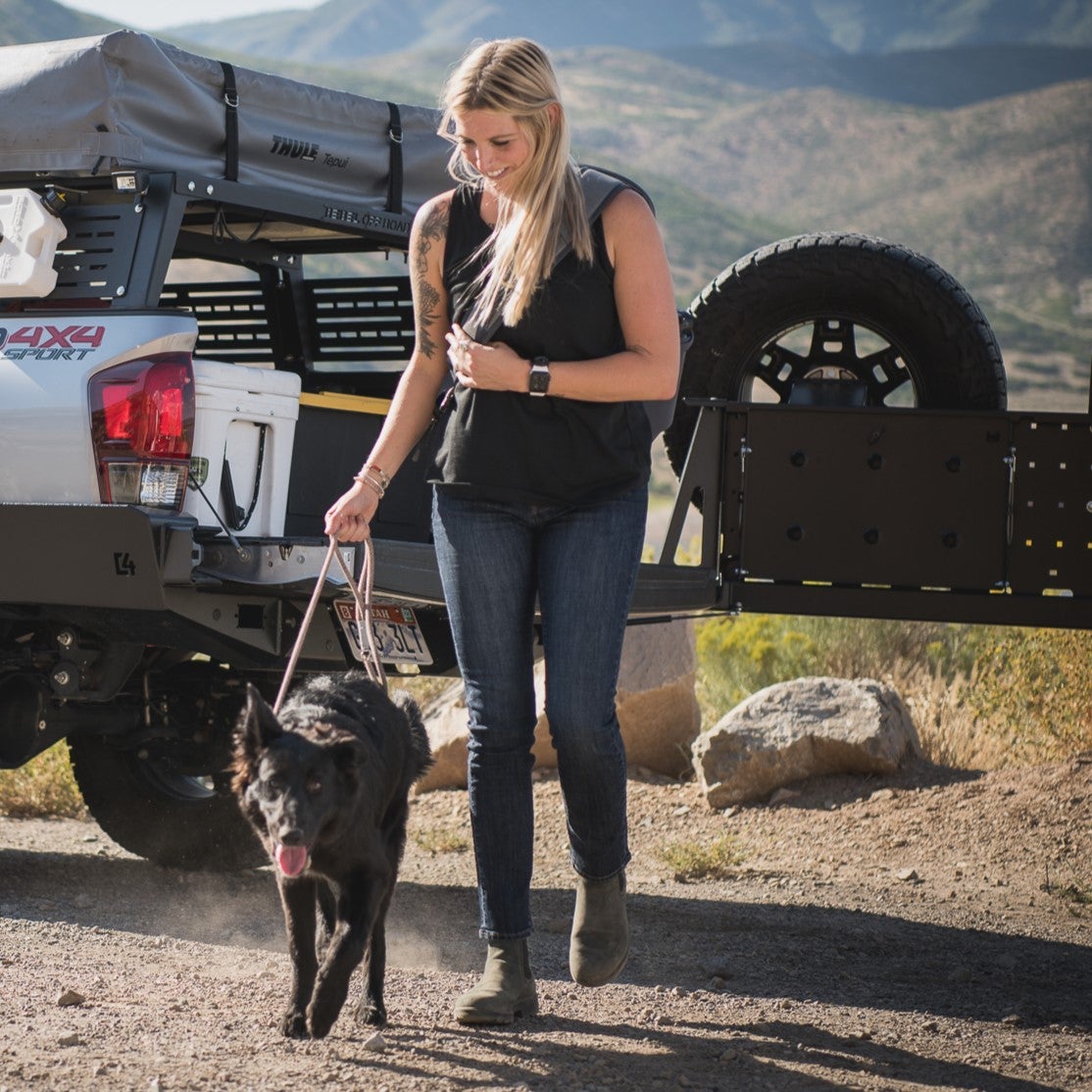 Photograph woman walking black dog near a Toyota Tacoma with Thule Tepui rooftop tent in a mountainous landscape featuring a spare tire and a gray, rocky, dusty trail