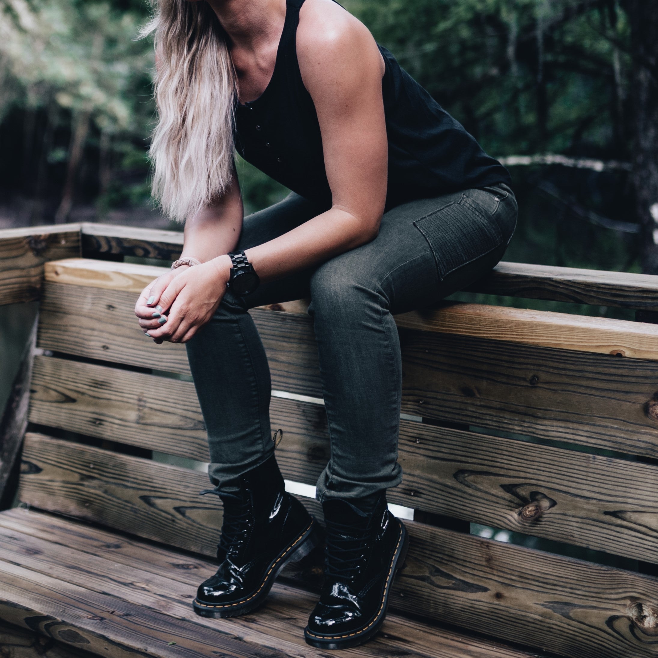 Photograph of a woman wearing black Doc Martens sitting on a rustic wooden bench in a forest with blonde hair and a black tank top