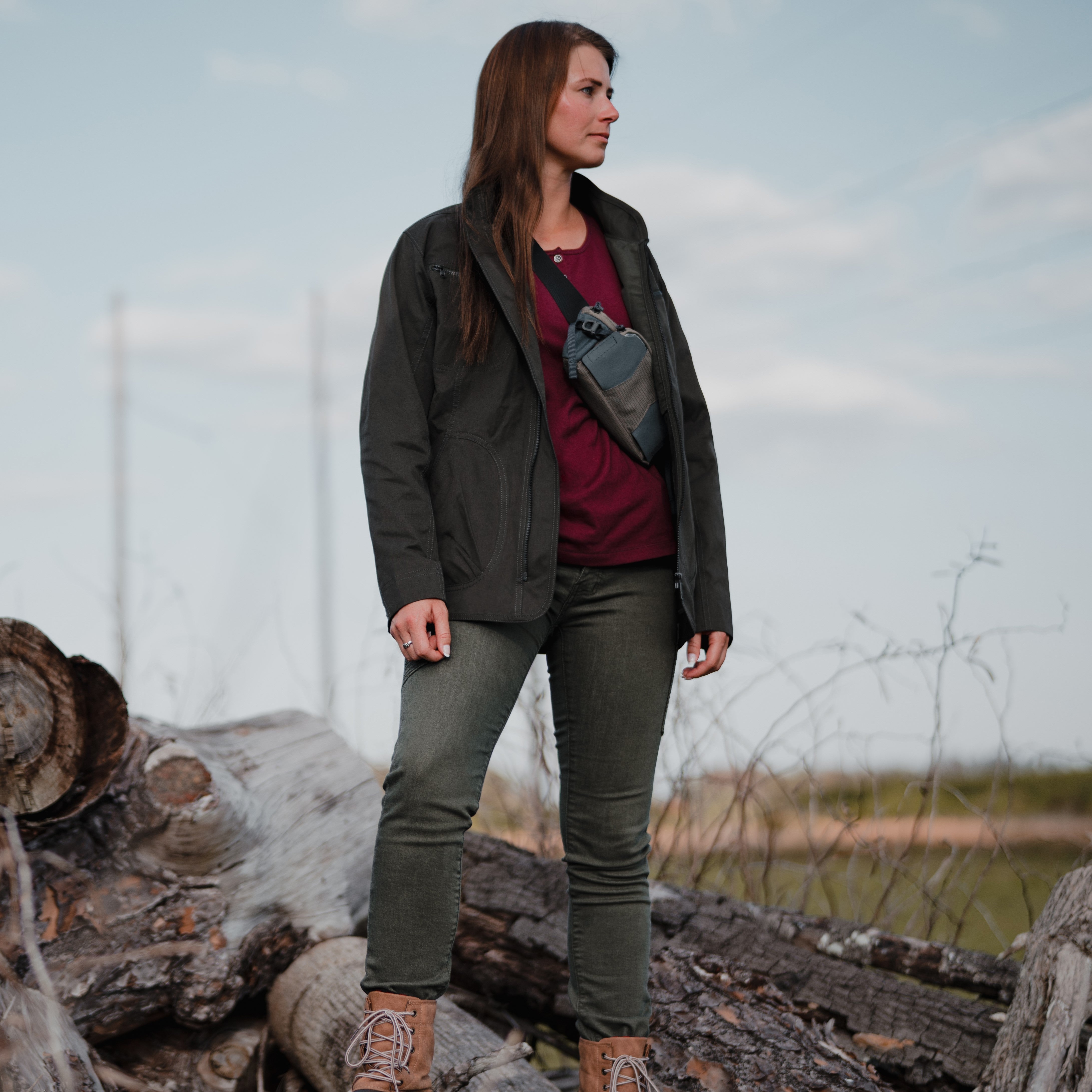 Photography a woman in olive green jacket and burgundy top standing on a pile of logs against a muted sky with distant power lines and dry brush showing off her tan boots and olive green pants