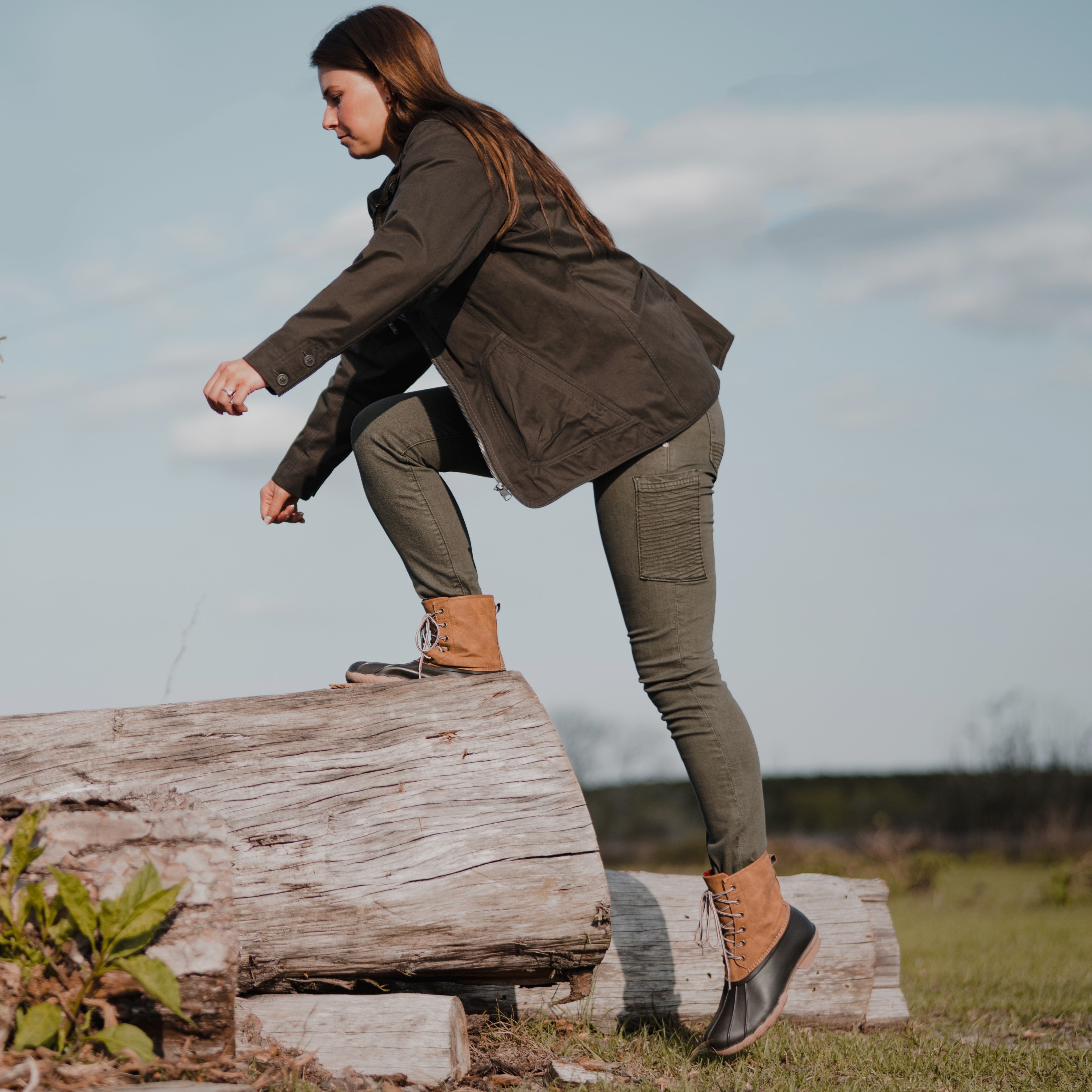Photograph of a woman in a olive green jacket and olive green pants stepping onto a large grey log in a grassy field with tan and black duck boots