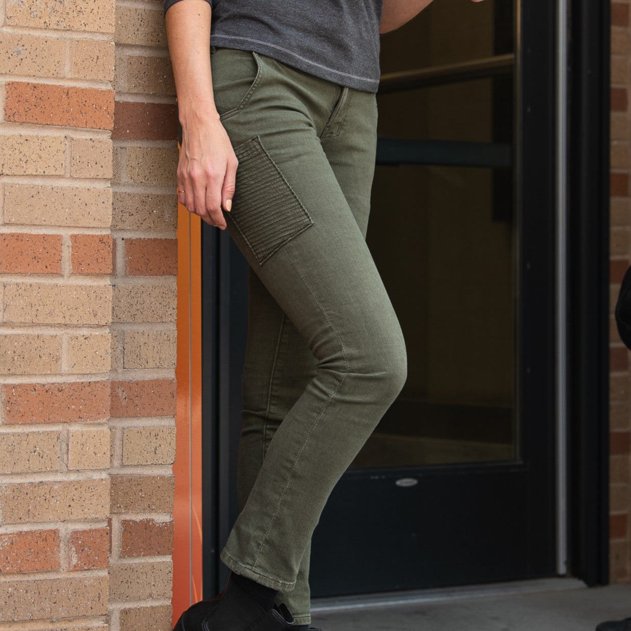 Photograph of olive green women's jeans with a unique moto-inspired detail near a brick wall and a dark door featuring beige and reddish-brown brick tones and subtle texture