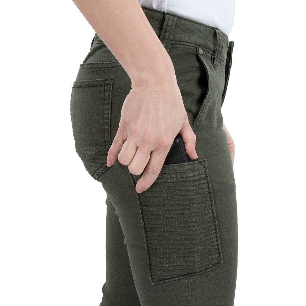 Product shot showing a woman's hand placing a black handgun magazine into a dark olive green pants pocket featuring reinforced stitching and a unique rear yoke design