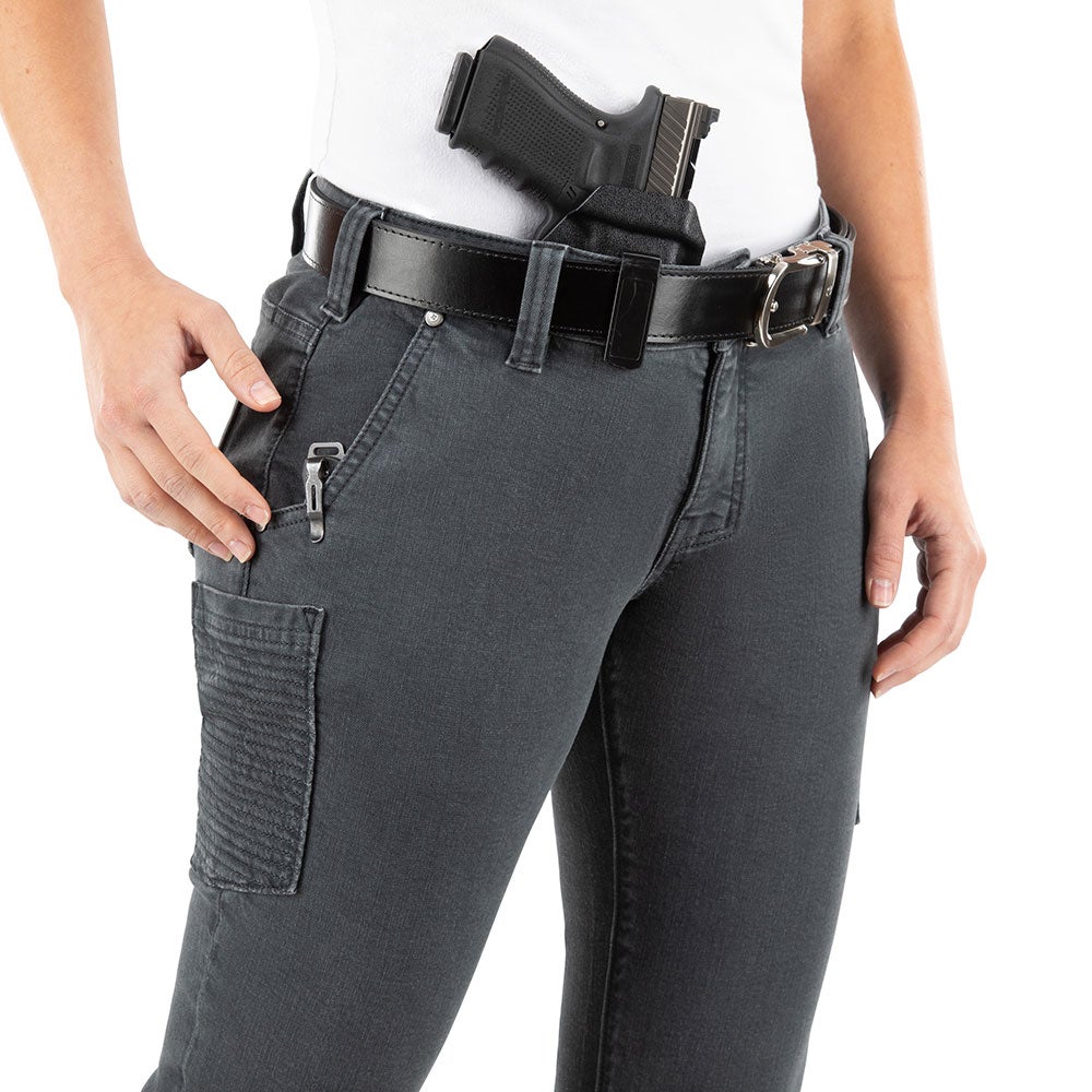 Product photography shows a woman wearing dark gray jeans with a concealed firearm a black leather belt and a silver pocket knife the jeans have reinforced stitching and multiple pockets