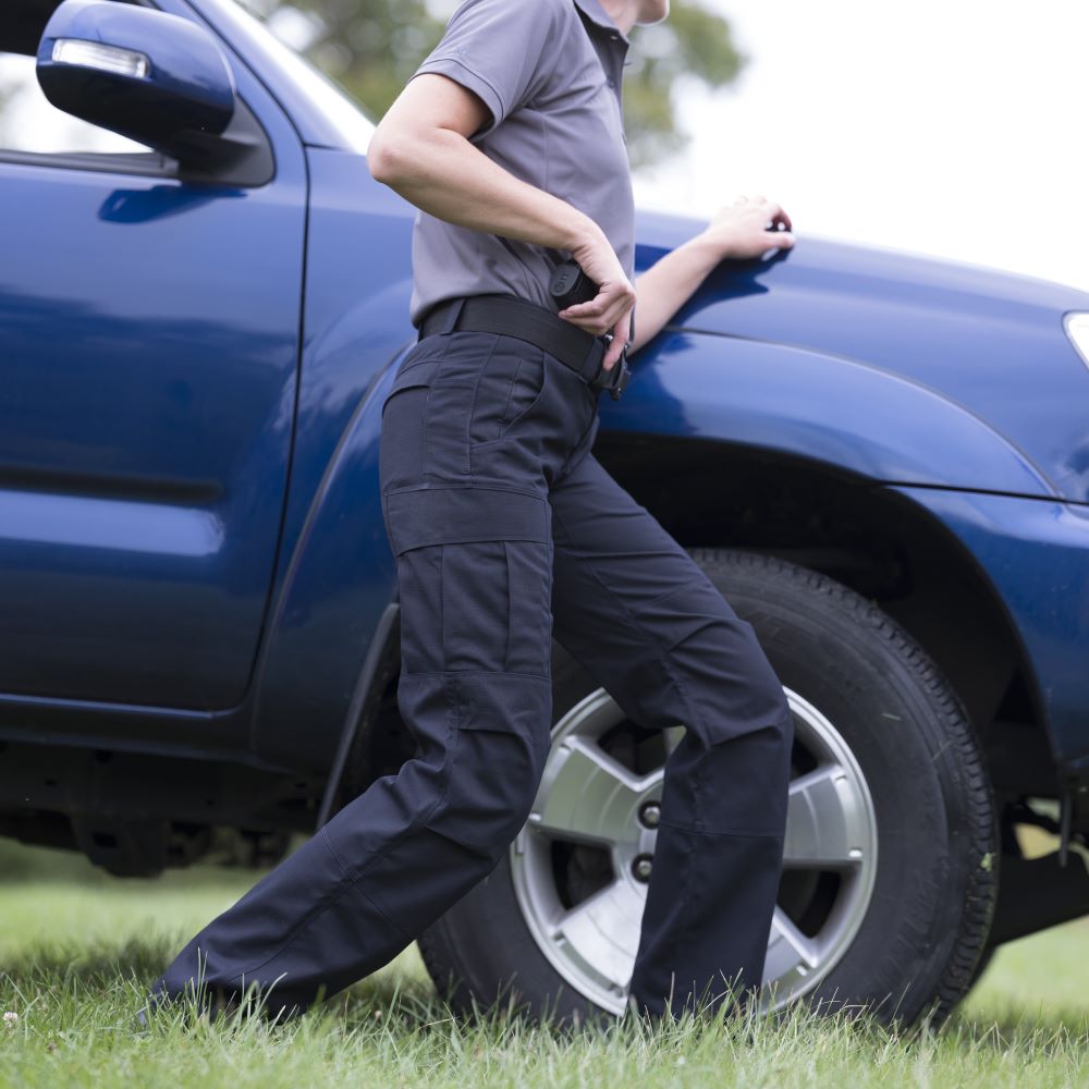 Photograph of a woman in dark tactical pants and a gray shirt next to a blue truck outdoors featuring a silver wheel cargo pockets and a holstered firearm