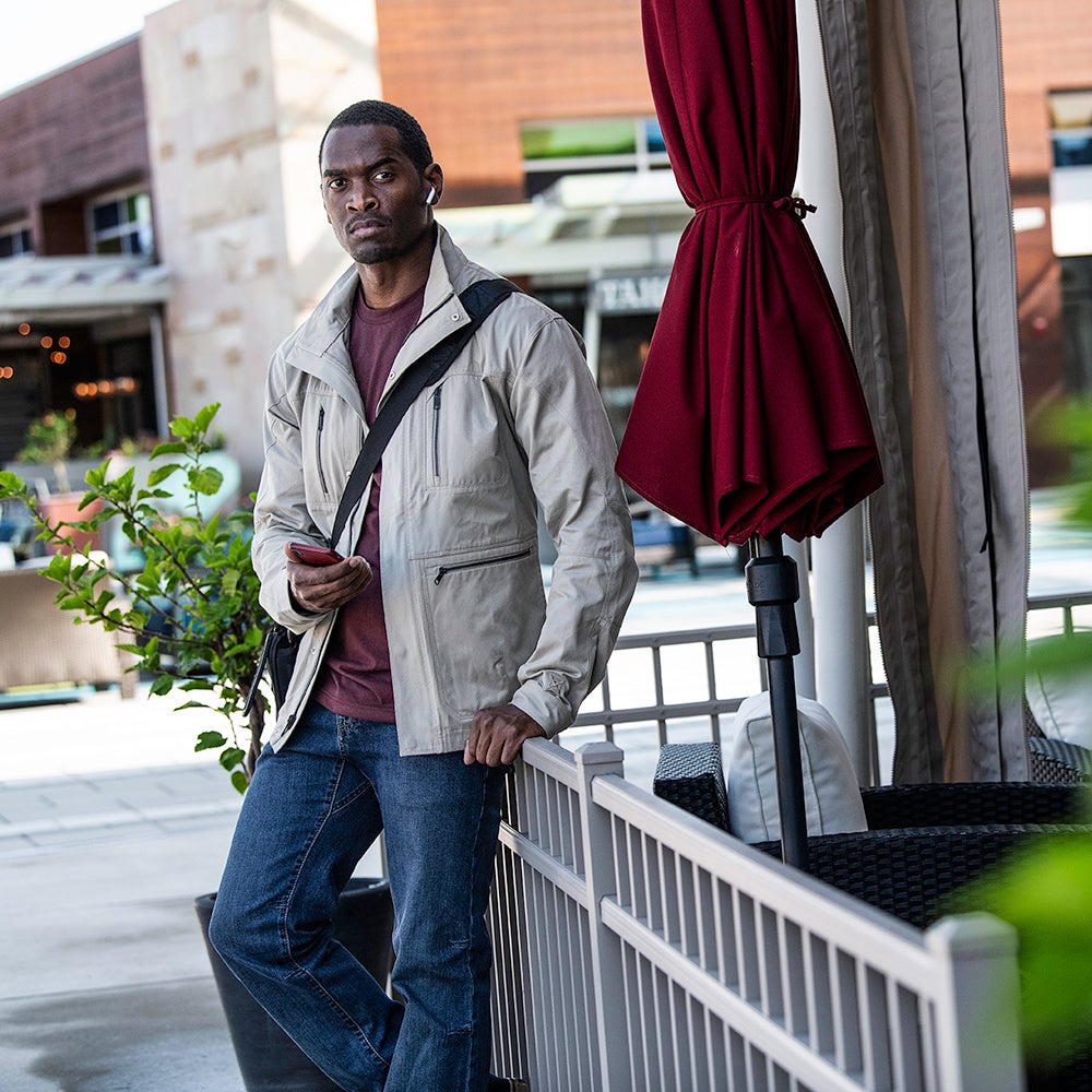 Photograph of a man in a beige jacket and blue jeans holding a phone outside a building with a maroon umbrella, gray fence, and lush green plants.