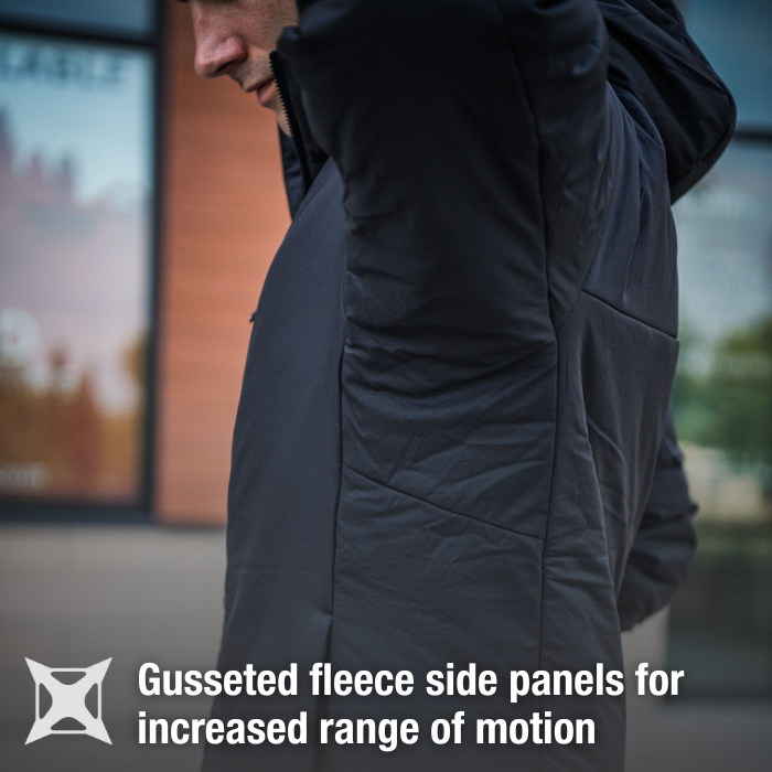 Product photography showing a man wearing a black fleece jacket with gusset detailing and a textured surface outdoors near a modern building with large windows