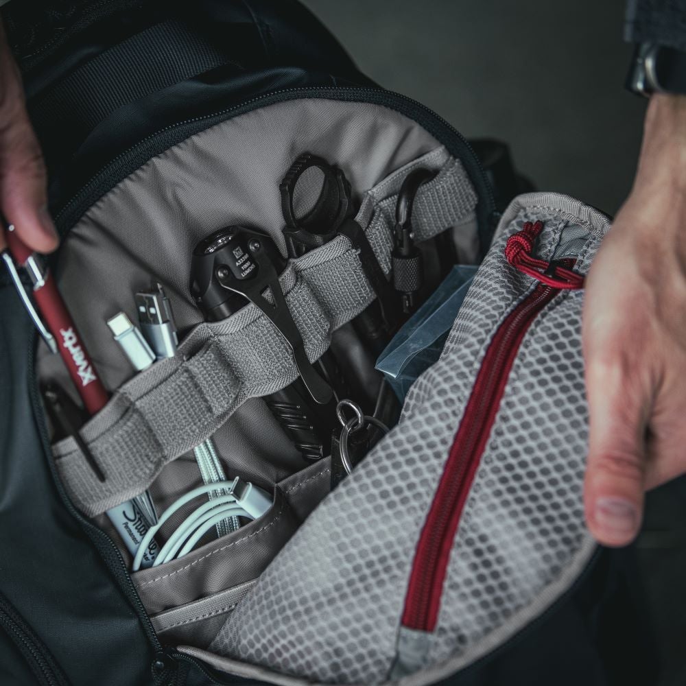 Photograph of a well-organized backpack interior showing various tools, a red pen, USB cables, and a flashlight with a red zipper pull and grey mesh pocket