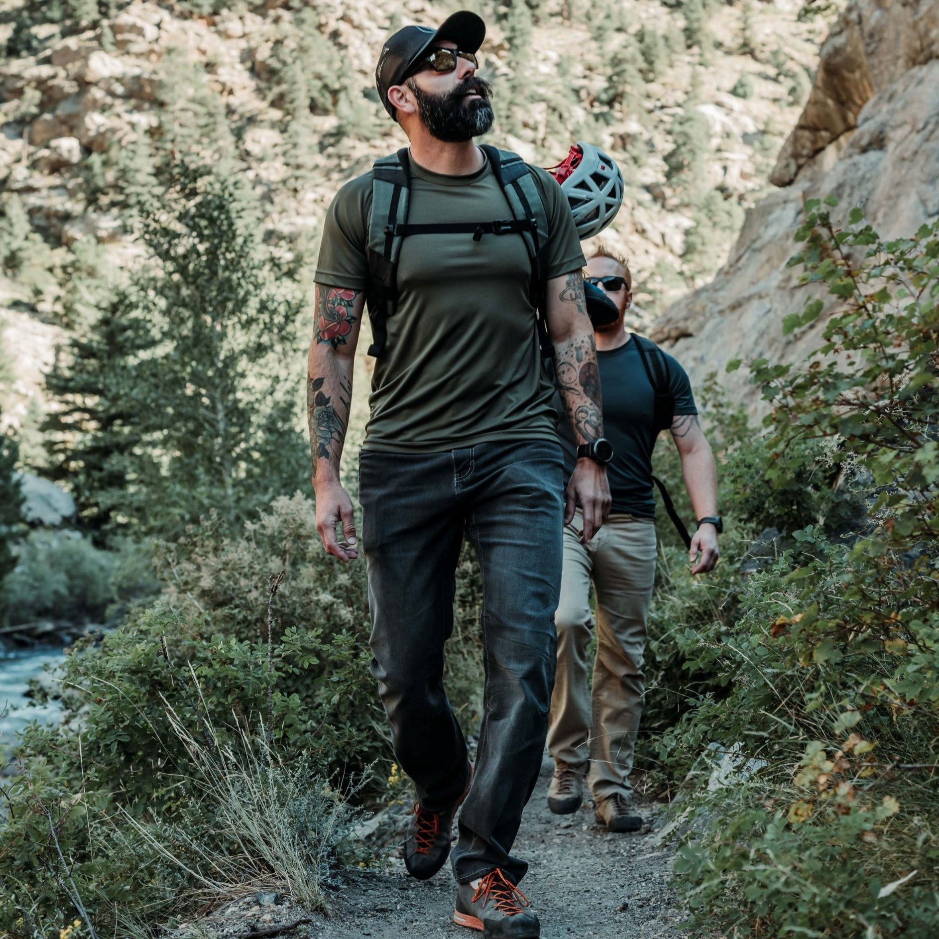Photograph of two men hiking a trail near a river with rocky cliffs and lush green vegetation featuring a man with tattoos olive shirt and orange shoelaces