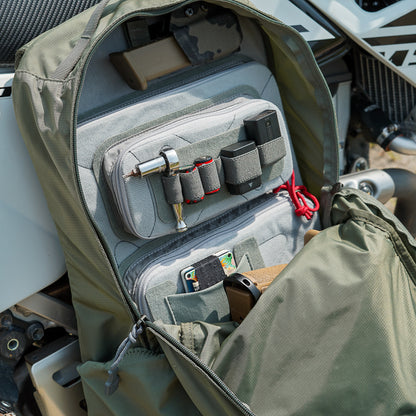 Photograph of an olive-green backpack's organized interior showing various tools, a firearm, and gray pouches next to a motorcycle
